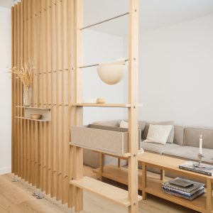 WoodUpp-rumsavskiljare i ljus ek med vertikala lameller, metallstänger och integrerade hyllplan med naturlig oljad yta. Bilden visar WoodUpp-hyllor, en utdragbar tyglåda/box och en hängande rund kruka på stång, samt en liten väggmonterad hylla med vas — stilren och funktionell i vardagsrummet.