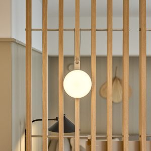 Bild av WoodUpp:s vertikala eklameller som bildar en rumsavdelare med horisontella metallstänger och infästa mässingsfärgade clips. En rund, vithängande lampa hänger i ett WoodUpp-krokfäste mitt i spjälorna, medan en svart bordslampa syns i bakgrunden bakom lamellerna i ljus ekfinish och minimalistisk design.