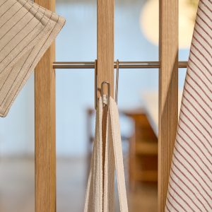 Närbild av WoodUpps träavskiljare i ljus ek med en tunn rostfri metallstång och två WoodUpp S‑krokar som håller upp textilier: en randig kökshandduk, en tygvante och ett naturfärgat bomullsband. Bilden framhäver träådring, metallfinish, mjuka tygtexturer, varm bakgrundsbelysning i skandinavisk stil.