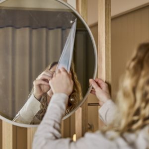 Person tar bort skyddsfilm från en rund spegel från WoodUpp med tunn grå ram, monterad på ljusa träpaneler med vertikala lister från WoodUpp. Bilden visar spegelns reflektiv yta, trästommens synliga ådring och händer som fäster spegeln mot väggstommen i varm inomhusmiljö med dov belysning.