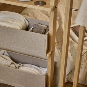 WoodUpps öppna klädställning i ljus ek med två staplade beige textilboxar fyllda med hopvikta tröjor och strumpor, synliga trästolpar, metallfästen och en tunn garderobsstång. Bilden visar detaljer av material och funktion i ett neutralt, mjukt sovrumsarrangemang med filtklädda sidoväggar och varma naturtoner.
