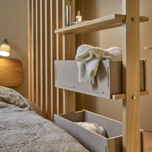 WoodUpps massiva ram i ljus ek visar en smal rumsavdelare med hyllplan och två grå filtboxar från WoodUpp, fyllda med vikta stickade plagg. Bilden visar även sängkant, lamellvägg och en liten hylla med vas och glasflaska i ett varmt, dämpat sovrumsljus.