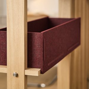 WoodUpp hyllställ i ljus ek med synlig sexkantsbult och liten hyllkonsol, där en mörkröd filtlåda från WoodUpp sitter inskjuten. Bilden visar närbild av den kantade filtboxen i ullliknande material och trästommen med enkel, modulär konstruktion med diskreta infällda handtag och varm, matt yta, samt rostfri metallknapp.