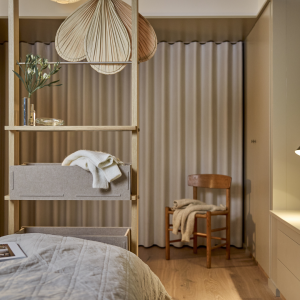 En mysig sovrumsmiljö med sängram och smarta, hängande sängbord i ljust trä från WoodUpp. Sängborden har lådor i grå filt och är dekorerade med en vas och en tröja. Sängen är bäddad med ett grått, quiltat överkast. I bakgrunden syns en trästol och gardiner.