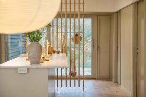 Bild av ett ljust köksrum med WoodUpps golv‑till‑tak‑spjälor i trä som rumsavdelare, med horisontella stag och hängande handduk samt nätpåse. I förgrunden syns en köksö med vas, tända ljus och ljust trägolv, stora glasdörrar mot trädgården.