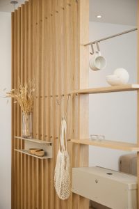 WoodUpps ljust träskärm med vertikala ribbor och inbyggda hyllor visas, kompletterad av beige metallhylla, hängkrokar och en avtagbar förvaringsbox. Bilden visar även mugg på skena, nätkasse, glasvas med torkade gräs, två små träskålar, snäckformad keramik och tända ljus i varma toner.