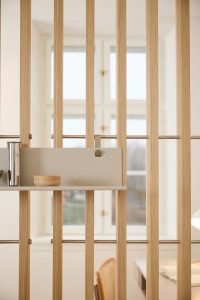 Bild av WoodUpps luftiga rumsavskiljare i ljust ek med vertikala lameller och tunna rostfria tvärstag. På lamellerna sitter en beige WoodUpp hylla/organiseringspanel med metallfäste och logo, med en liten träskål och rostfri behållare placerade på hyllplanet i ett ljust, minimalistiskt kontorsrum med fönster i bakgrunden.