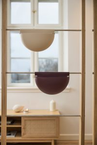 Bild visar WoodUpp-produkter: två hängande halvcirkelformade förvaringskärl/krukor i beige och mörklila som hänger på rostfria metallstänger i ett ljust träramverk. Släta, minimalistiska ytor i matt finish, praktiska för förvaring eller växter, placerade framför ett fönster i skandinavisk stil.