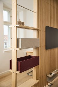Bild visar WoodUpps modulära hyll- och rumsavdelare i ljus ek med synliga bultfästen, två infällbara filtbackar — en grå och en vinröd — på träskenor, en liten hylla med garnnystan, framför slät träpanel och väggmonterad TV, praktisk förvaring och avskärmning i skandinavisk stil.