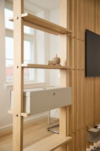 WoodUpps ljusa träavdelare med öppna hyllplan visas i bild, monterad i ekfinish med synliga metallsprintar. På hyllan sitter en grå-beige WoodUpp kabelbox med ventilationshål och slits för kablar. Dekoration med snöre och en TV på ribbad trävägg syns i bakgrunden.