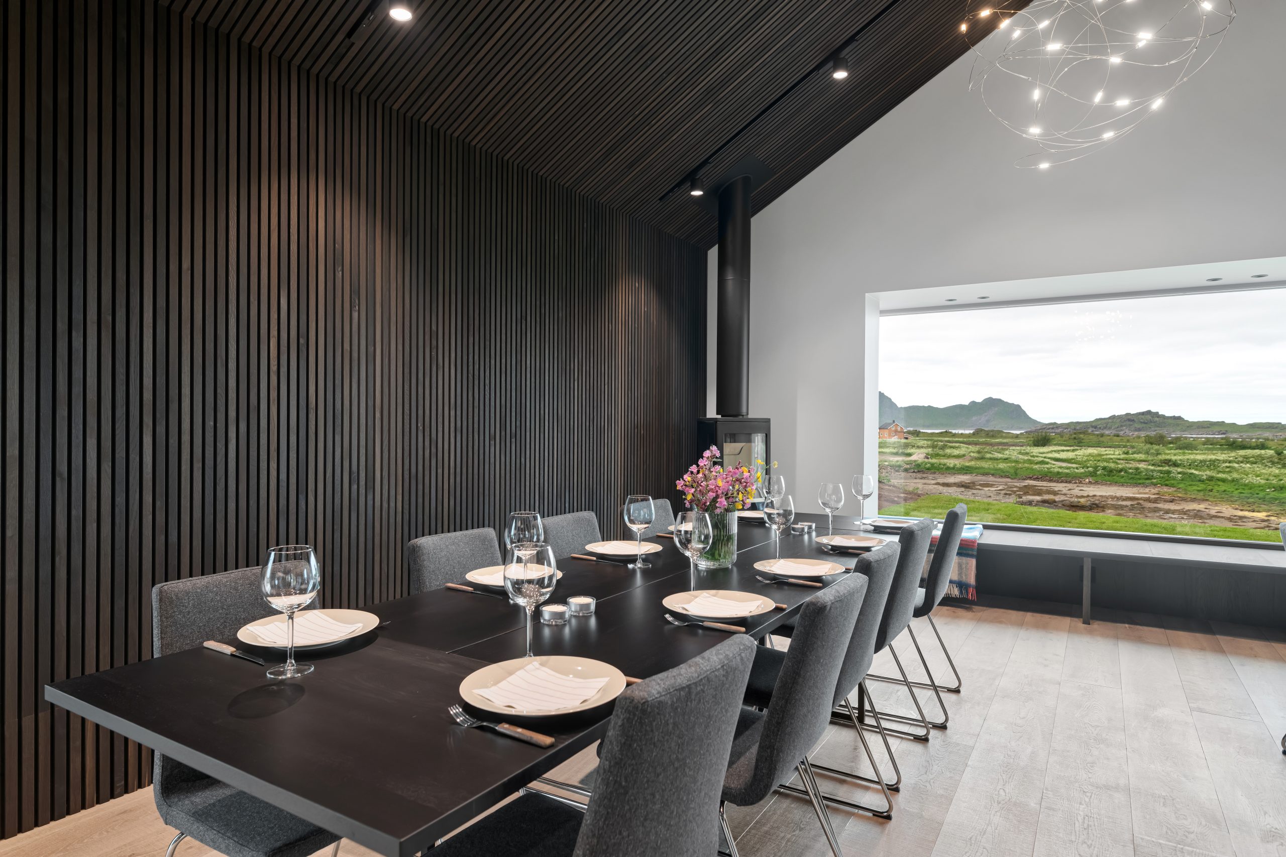 Matplats med WoodUpp vägg- och takpaneler i mörka, svartbetsade vertikala träslatsribb som skapar ett slimmat uttryck med djupa skuggor. Ett svart matbord är dukat med glas och porslin, omgiven av grå stoppade stolar och en vas med blommor. Stort fönster visar grönt kustlandskap.