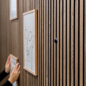 WoodUpp trälamellvägg i ek med smala vertikala spalter, där en svart WoodUpp magnetisk bildhängare håller upp ramar. Bilden visar två enkla träramar hängande, en hand som justerar en ram och detaljer av slät trästruktur och magnetfäste.